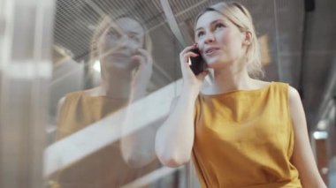Güzel sarışın iş kadını, şık sarı elbiseli, ofisteki cam duvara yaslanırken gülümseyen ve cep telefonuyla konuşan.