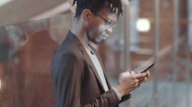 Gözlüklü Afro-Amerikan genç bir adamın yan görüntüsü ve cam duvara yaslanmış şık bir kıyafet ve ofiste çalışırken akıllı telefondan mesajlaşması.