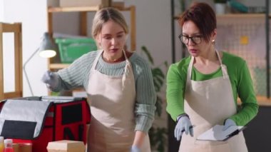 Önlüklü ve tek kullanımlık eldivenli bir kadın, elinde not panosu olan ve teslimat için yiyecek paketlerini torbaya koyarken genç bayan meslektaşına talimat veren bir kadın.