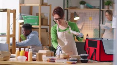 Beyaz önlüklü, eldivenli orta yaşlı bir kadın teslimat için eko dostu yiyecek kutularını paketliyor ve ticari mutfakta çalışırken notlar alıyor.