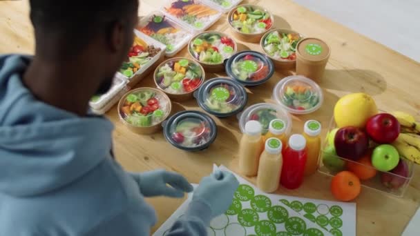 在健康的送餐服务中 戴着手套的黑人男子在装有有机食品的容器上贴上贴纸 从肩膀上方高角度拍了照 — 图库视频影像