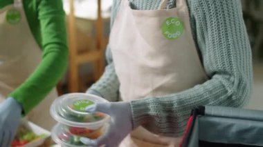 Önlüklü, eldivenli ve yüz maskeli genç bir kadının Coronavirus salgını sırasında sağlıklı yemek dağıtım servisinde çalışırken iş arkadaşının yardımıyla çantasına yemek paketlediği el kamerası görüntüleri.