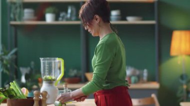 Beyaz bir kadının taze salatalık keserken ve evdeki mutfak masasında sağlıklı yemek pişirirken parçaları miksere koyarkenki görüntüsü.