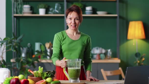 白种人女人在拍摄健康饮食博客的同时 从搅拌器罐子向杯子里倒出绿色的冰沙 看着相机 告诉菜谱 — 图库视频影像