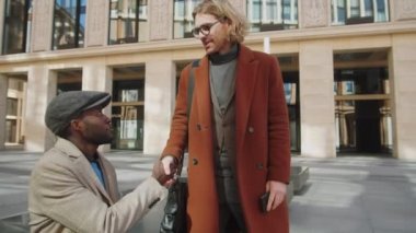 Genç beyaz iş adamı, dışarıda dizüstü bilgisayar kullanırken Afro-Amerikalı iş arkadaşına yürüyor, onu el sıkışarak selamlıyor, konuşuyor ve akıllı telefon ekranında bir şeyler gösteriyor.