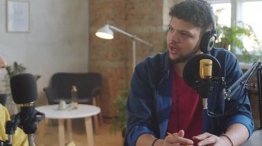 Kulaklıklı genç adam podcast stüdyosunda bayan konukla röportaj yaparken mikrofonla konuşuyor.