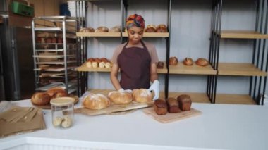 Önlüklü, eldivenli, fırında somun ekmeği olan ve kameraya bakıp gülümseyen güzel Afro-Amerikan kadının portresini büyüt.