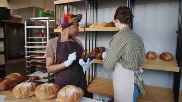 若いアフリカ系アメリカ人女性がデジタルタブレットで作業し パン屋の木のディスプレイラックに新鮮なパンを置くとして白人女性の同僚と話をする — ストック動画