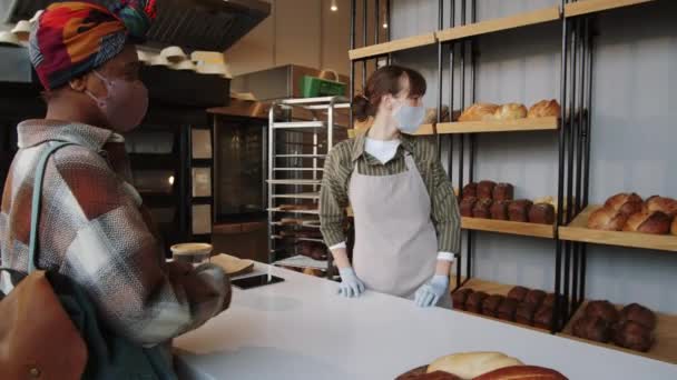 アフリカ系アメリカ人女性のための紙袋にパンのエプロン マスク 手袋包装のローフの白人のセールスマンは Covid 19流行中にパン屋で働いている間 — ストック動画