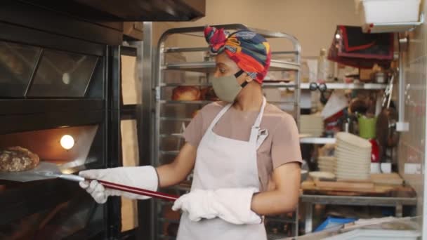 顔のマスク 保護手袋 エプロンをしたアフリカ系アメリカ人女性のパン屋は コビト19のパンデミックの間にパン屋で働いている間 シャベルでオーブンから焼きたてのパンを取り出し 棚にパンを置く — ストック動画
