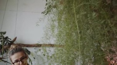 Genç ve çekici bir kadın, gözlükleriyle kuşkonmaz eğreltiotu sıkarken kapalı bahçedeki bitkilerle ilgileniyor.