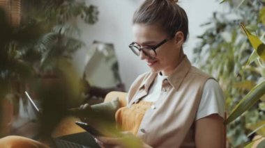 Gözlüklü ve günlük kıyafetli genç bir kadın akıllı telefonuyla selfie çekiyor ve ev bahçesinde bitkilerle dolu dizüstü bilgisayarıyla oturup gülüyor.