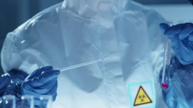 Laboratuvarda antiviral ilaç araştırması yaparken koruyucu giysili, gözlüklü, maskeli ve eldivenli bilim adamının Coronavirus yazıtlı kırmızı sıvı maddeyi test tüpünde sallarken çekimini azalt.