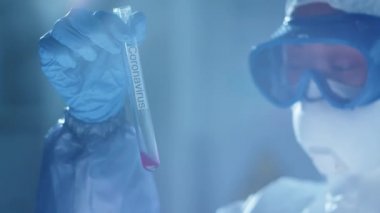 Koruyucu giysi, gözlük, maske ve eldiven giyen kadın bilim adamı laboratuarda antiviral tıp üzerinde çalışırken Coronavirus yazıtlarıyla test tüpünde sıvı maddeyi inceliyor.
