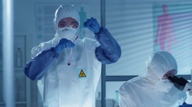 Koruyucu giysili, gözlüklü, maskeli ve eldivenli iki bilim adamı laboratuvarda antiviral ilaç üzerinde birlikte çalışıyorlar: kadın test tüpüne kan döküyor ve adam mikroskop kullanıyor.