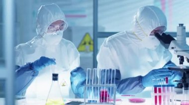 Koruyucu giysiler, gözlükler, maskeler ve eldivenler içinde antiviral tedavi üzerine araştırma yapan iki laboratuvar çalışanı: kadın bilim adamı test tüpüne kan döküyor ve erkek mikroskoptan bakıyor.