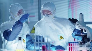 Koruyucu üniformalı, maskeli, gözlüklü ve eldivenli iki profesyonel bilim adamı laboratuarda antiviral tıp üzerinde çalışırken test tüpünde kırmızı maddeyi inceliyor ve tartışıyor.