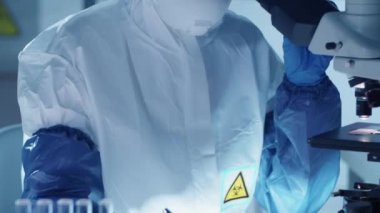 Koruyucu maskeli, takım elbiseli, eldiven ve gözlüklü kadın viroloji uzmanı mikroskop kullanıyor ve laboratuarda koronavirüs tedavisi üzerine araştırma yaparken panoya not alıyor.