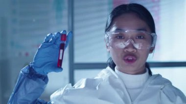 Korumacı kıyafetli, gözlüklü ve eldivenli Asyalı kadın bilim adamının göğüs görüntüsü. Test tüpünde üzerinde Covid-19 yazısıyla kan örneği tutuyor. Kameraya bakıyor ve bir şeyler söylüyor.