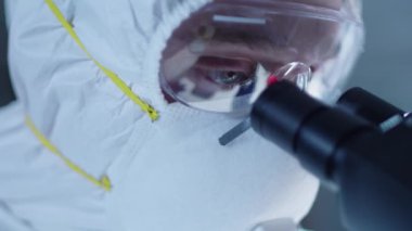 Koruyucu maskeli, gözlüklü ve takım elbiseli bir erkek bilim adamı yüzünü mikroskoba yaklaştırıyor ve laboratuarda antiviral tıp üzerinde çalışırken dürbünlerden bakıyor. Covid-19 salgın konsepti
