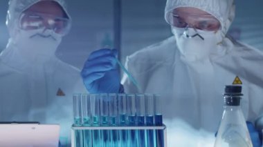 Koruyucu takım elbiseli, eldivenli, gözlüklü ve maskeli bir bilim adamı deney tüpüne mavi sıvıyı damlatırken, asistanı da panoya notlar yazıyor. Covid-19 salgın konsepti