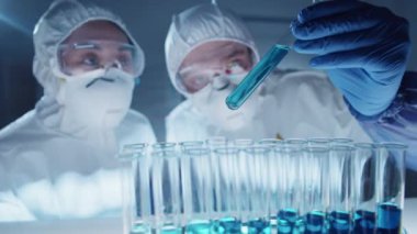 Laboratuvarda antiviral aşı üzerinde çalışırken test tüpünde mavi sıvıyı inceleyen ve tartışan koruyucu ekipmanlı kadın ve erkek çok ırklı virologların odaklanmış görüntüleri.