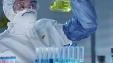 Koruyucu üniformalı, maskeli, gözlüklü ve eldivenli kadın Asyalı bilim adamının laboratuarda antiviral aşı üzerinde çalışırken cam şişede sarı sıvıyı incelerkenki görüntülerini yukarı kaldır. Covid-19 salgın konsepti