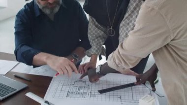 Çok ırklı erkek ve kadın mimarlardan oluşan bir ekibin ofis masasında birlikte çalışırken kat planını tartışırken yüksek açılı yakın plan çekimi.