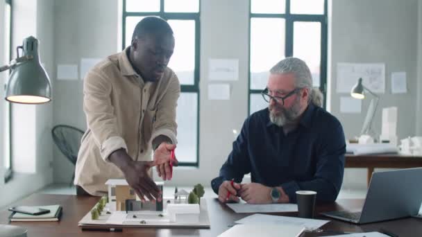 若い黒人男性建築家は オフィスで一緒にプロジェクトに取り組んでいる間 シニア同僚と建築モデルを議論 — ストック動画
