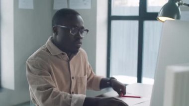 Genç siyah erkek mimar gözlüklü ve günlük kıyafetleriyle bilgisayarda yazı yazıyor ve ofiste çalışırken notlar alıyor.