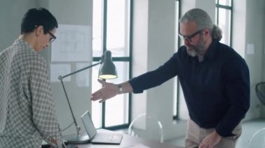 Gri saçlı ve sakallı yaşlı bir erkek mimarın kadın meslektaşıyla el sıkışırken ve onunla ofis masasında konuşurken.