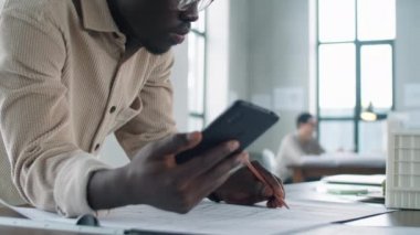 Genç Afro-Amerikan erkek mimarın akıllı telefon ekranına baktığı ve ofiste çalışırken zemini kalemle çizdiği el kamerası görüntüsü.