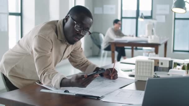 アフリカ系アメリカ人の若い男性建築家がオフィステーブルにもたれかかり スマートフォンを持ち 鉛筆でフロアプランを描き カメラを見る — ストック動画