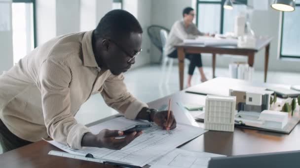 年轻的非洲裔美国男性建筑师靠在写字台上 用智能手机和铅笔在纸上画出平面图的弧形照片 — 图库视频影像