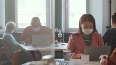 Coronavirus salgını sırasında meslektaşlarıyla birlikte açık uzay ofisinde çalışırken masasında oturup dizüstü bilgisayarda yazan koruyucu maskeli melez bir kadın.