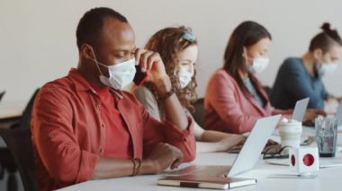Korunaklı yüz maskesi takan Afrikalı Amerikalı işadamı Coronavirus salgını sırasında açık uzay ofisindeki meslektaşlarıyla çalışırken cep telefonuyla konuşuyor ve masadaki bilgisayarını kullanıyor.
