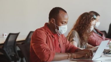 Genç Afro-Amerikalı iş adamı koruyucu yüz maskesi takıyor ve Coronavirus salgını sırasında çalışma masasında çalışırken kameraya bakıyor.