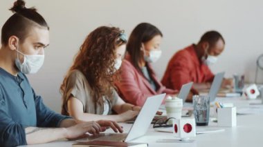 Koruyucu yüz maskesi takmış genç beyaz bir adam dizüstü bilgisayar kullanıyor ve Coronavirus salgını sırasında ofis masasında çok ırklı meslektaşlarıyla çalışırken kameraya poz veriyor.