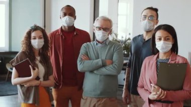 Korunaklı yüz maskeleri ve günlük kıyafetlerle karışık yaştaki kadın ve erkeklerden oluşan bir iş ekibi ofiste yan yana duruyor ve Coronavirus salgını sırasında kameraya bakıyor.