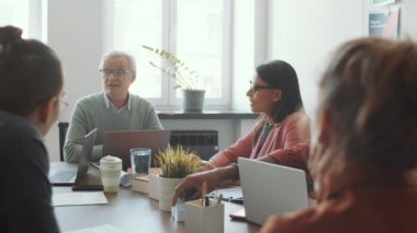 Üst düzey beyaz adamın toplantı masasında dizüstü bilgisayarıyla otururken ve ofiste iş projesinde birlikte çalışırken erkek ve kadın profesyonellerden oluşan genç bir ekip ile konuşurken PAN çekimi.