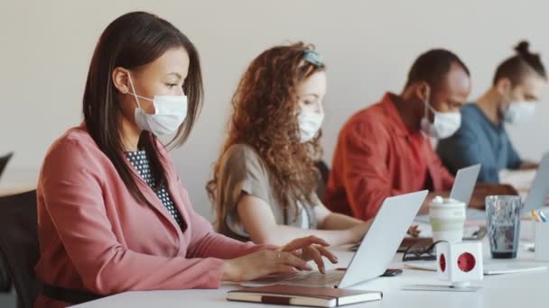デスクでラップトップを使用して顔のマスクで若い混合レースビジネス女性は Covid 19流行中に多様な同僚とオープンスペースオフィスで働いている間 — ストック動画