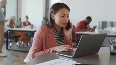 Güzel melez iş kadını gülümsüyor, cep telefonuyla konuşuyor ve açık uzay ofisinde çalışırken dizüstü bilgisayar kullanıyor.