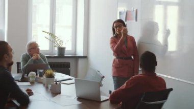 Güzel melez iş kadını beyaz tahtaya grafik çiziyor ve şirket toplantısında karışık yaştaki çok ırklı iş arkadaşlarına sunum yapıyor.