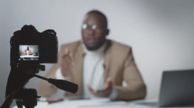 Stüdyoda oturan Afro-Amerikan erkek vlogcu tripodun üzerindeki dijital kameranın seçmeli odak noktası.