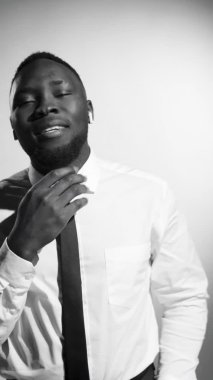 Resmi tişörtlü, kravatlı, gülümseyen, kameraya bakan ve stüdyoda poz verirken kablosuz kulaklıkla müziğin keyfini çıkaran Afro-Amerikan genç adamın siyah-beyaz dikey görüntüsü.