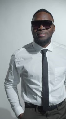 Resmi giyimli, modaya uygun güneş gözlüklü, gülümseyen ve stüdyoda kameraya poz veren Afro-Amerikan genç adamın dikey format görüntüleri.