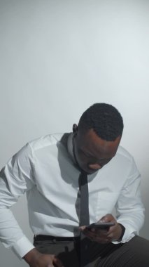 Atölyede beyaz arka planda oturan, akıllı telefon kullanan ve kameraya bakıp gülümseyen Afro-Amerikan gömlekli ve kravatlı genç adamın dikey format videosu.