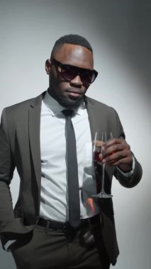 Zarif takım elbiseli, şık güneş gözlüklü, elinde şarap kadehi olan ve stüdyoda gülümseyerek kameraya poz veren genç Afro-Amerikan adamın dikey format görüntüleri.