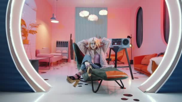 在粉红灯光的工作室里 穿着华丽服装的性感金发女人坐在艺术装饰扶手椅上 在老式盒式磁带唱机上与音乐共舞 放大了镜头 — 图库视频影像
