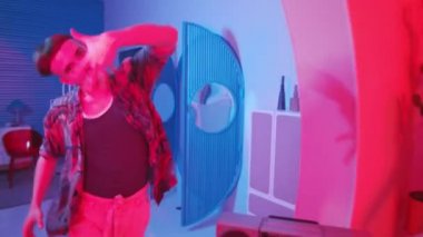 Genç hippi saç kesimli ve bıyıklı genç adam geçmişe dönük partiye giriyor ve arkadaşlarıyla birlikte neon ışıklı ve sisli havalı dans ediyor.